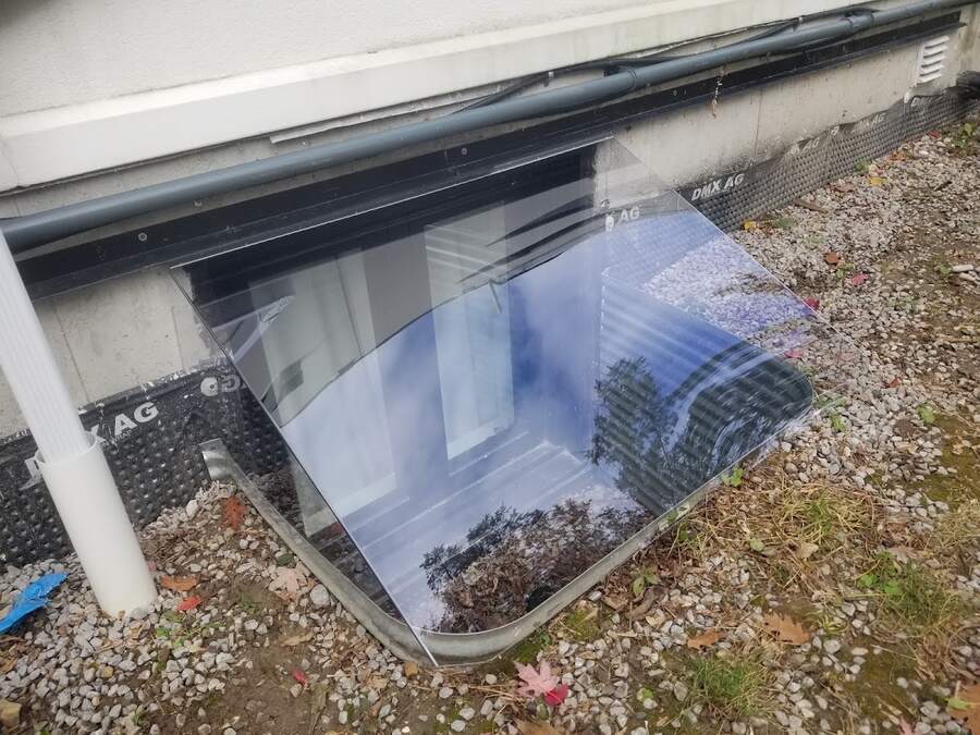 Clear custom window well cover fitted around exterior pipes